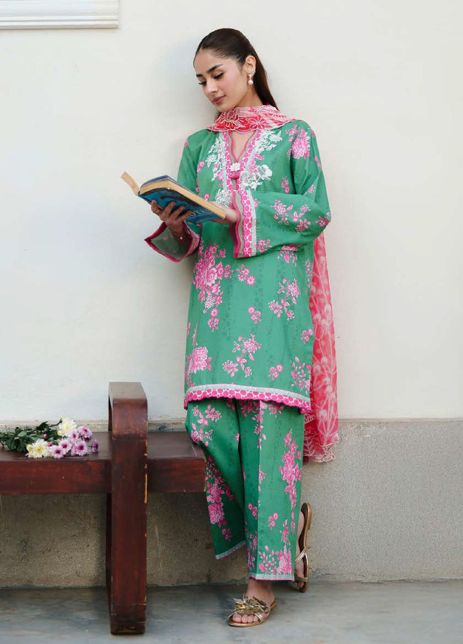 Woman reading a book dressed in a green embroidered floral lawn suit with pink accents and a matching pink chiffon dupatta, posing beside a wooden bench.