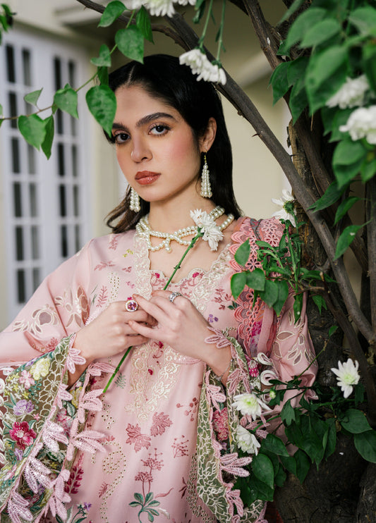 Close-up portrait of woman in a baby pink embroidered suit with pearl earrings and necklace holding a white flower, surrounded by greenery.