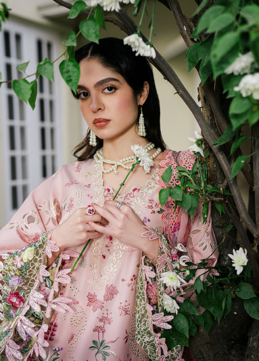Close-up portrait of woman in a baby pink embroidered suit with pearl earrings and necklace holding a white flower, surrounded by greenery.