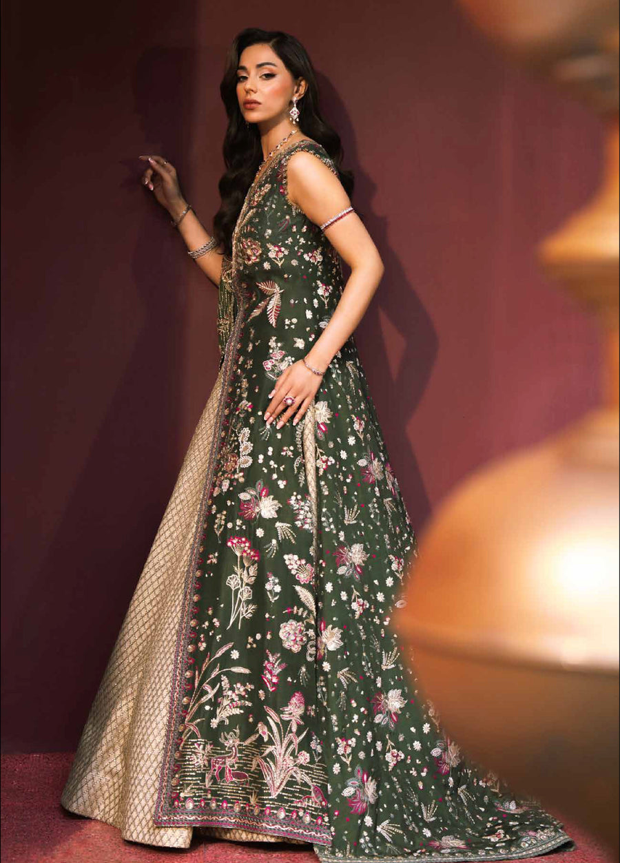 Side angle view of the woman facing left, highlighting the richly embroidered dark green tunic and golden lehenga with jewelry set and arm accessories, with blurred golden pillars in foreground.