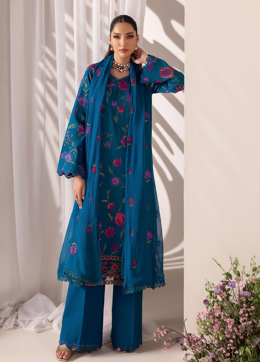 Woman in a blue embroidered lawn suit with matching chiffon dupatta draped over shoulders, standing near sheer white curtains and flowers
