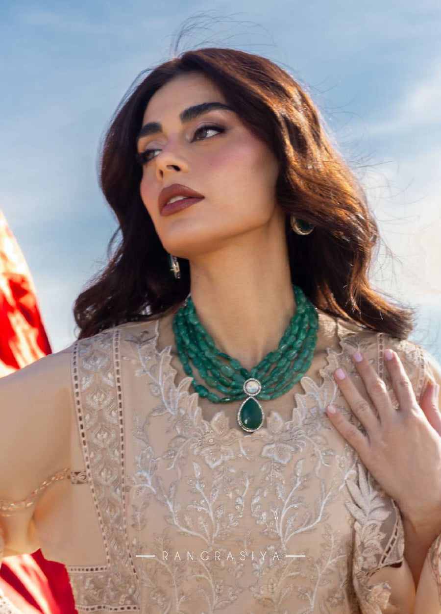 Portrait of woman with layered green stone necklace and earrings wearing beige embroidered lawn suit, showcasing embroidered neckline detail