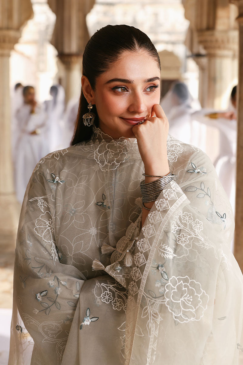 Woman smiling and looking away while wearing a beige embroidered lawn suit with organza dupatta draped over both shoulders in a courtyard setting