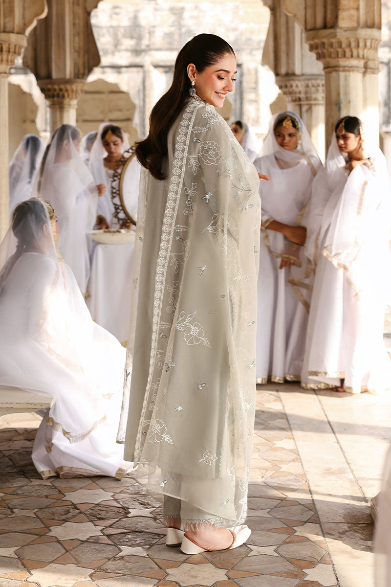 Back view of woman wearing beige embroidered lawn suit with sheer embroidered dupatta, standing in a traditional courtyard with women in white dresses behind her
