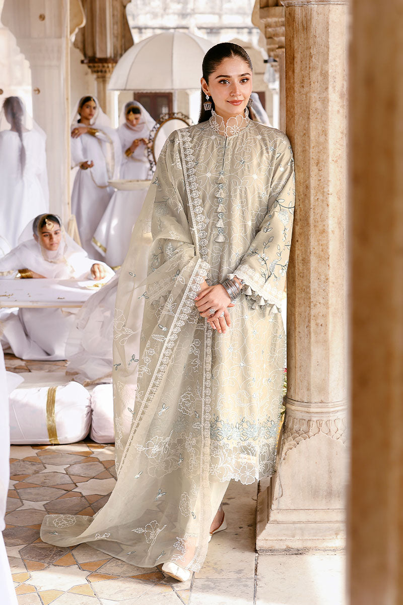 Woman wearing a beige embroidered lawn suit with floral and geometric patterns standing by a column in a traditional courtyard with women in white dresses in the background