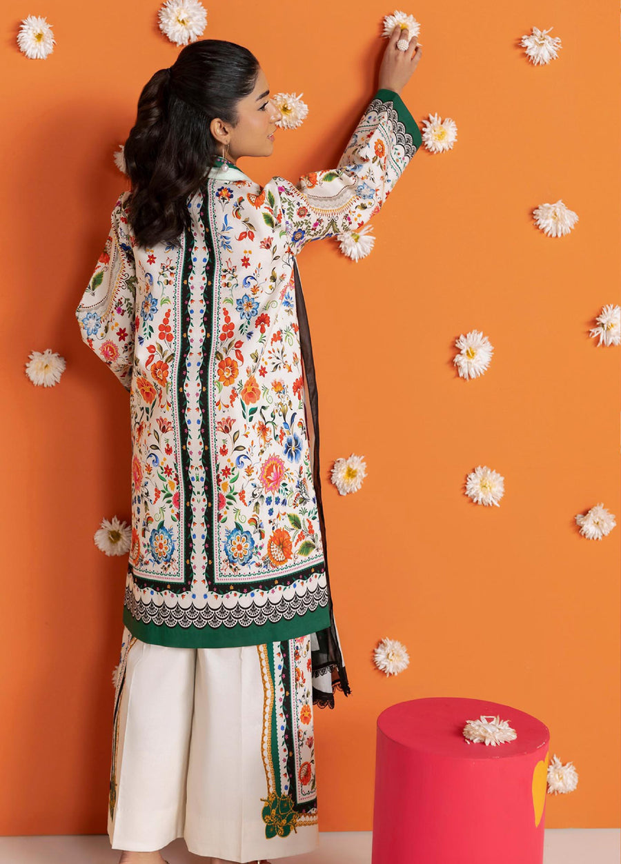 Rear view of woman touching a wall decorated with white flowers, displaying the back floral and lace-inspired print on the lawn shirt and trousers of the 3-piece suit.