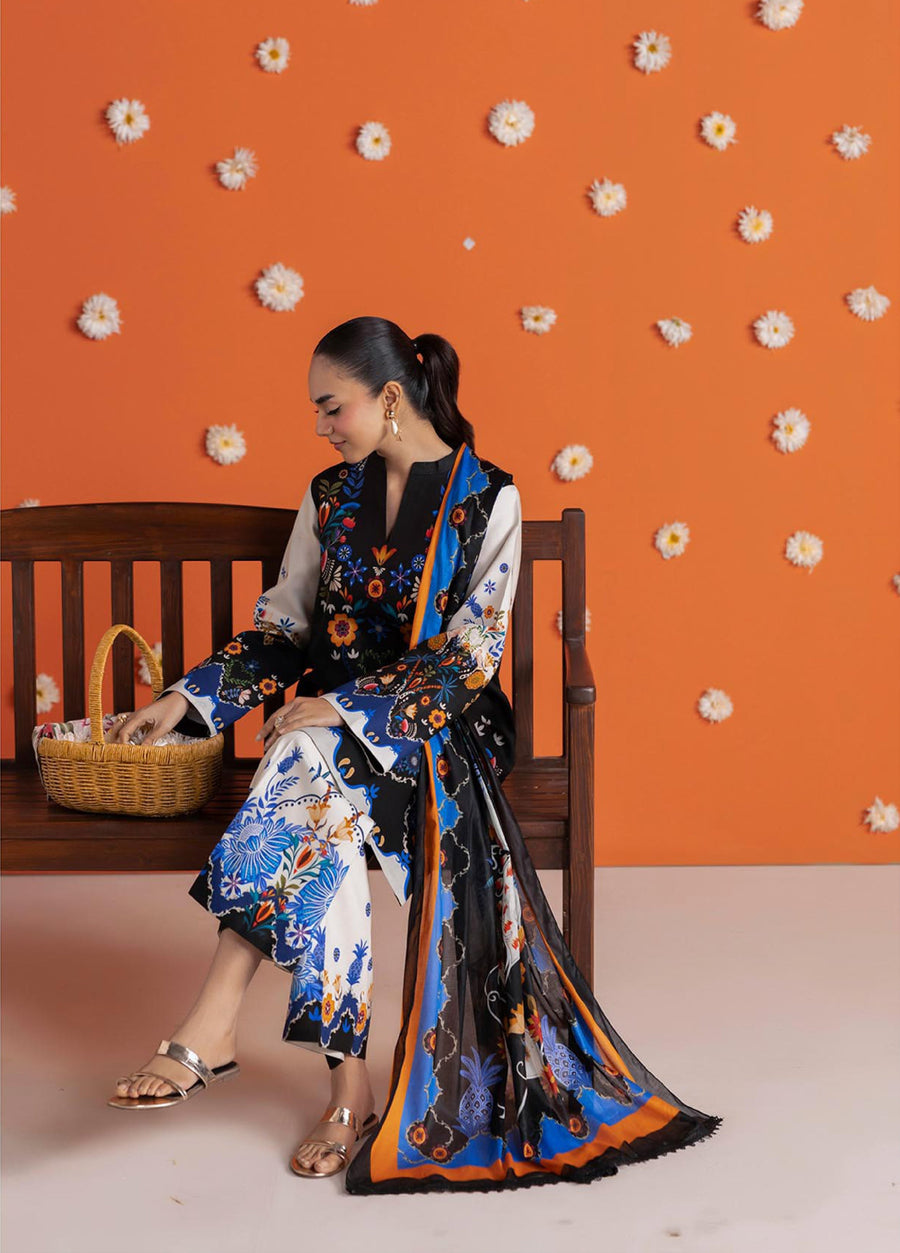 Woman sitting on a bench wearing a black printed lawn shirt, cream wide-leg trousers with blue floral designs, and a matching dupatta with floral patterns, orange wall background