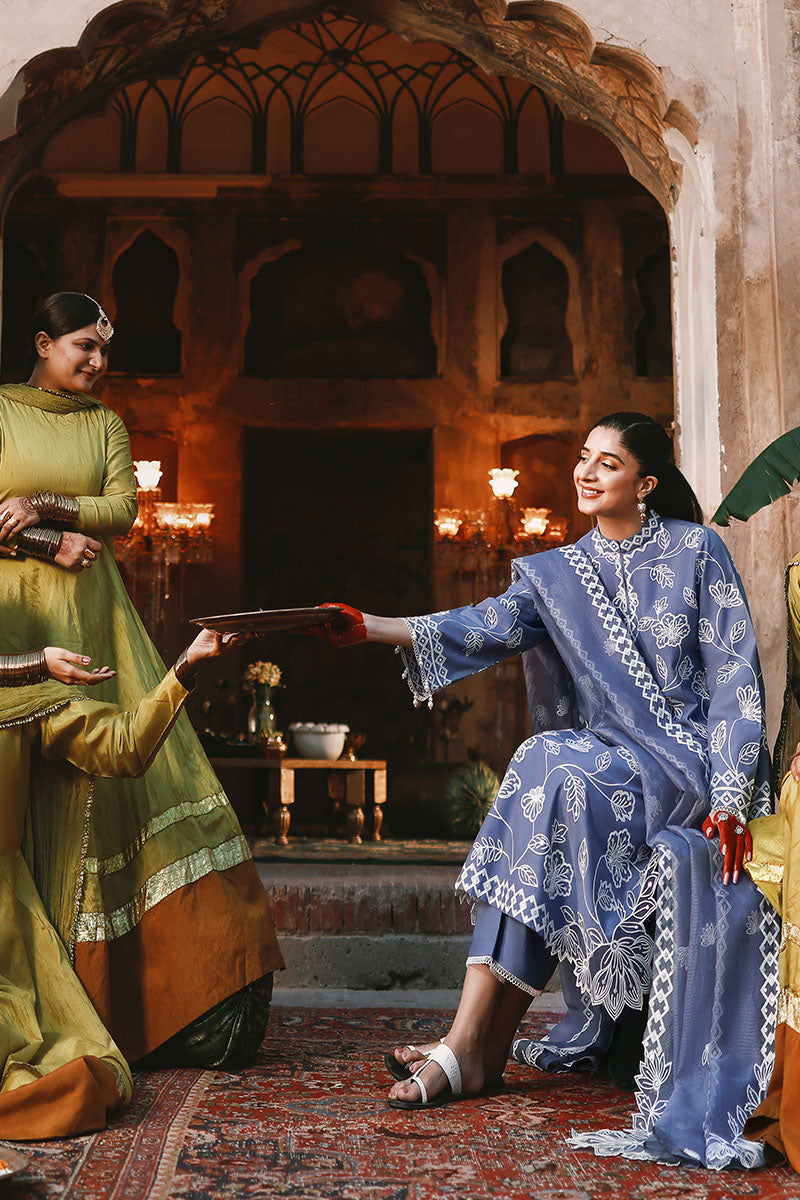 Woman seated wearing the blue embroidered 3-piece suit offering a tray, surrounded by women in green traditional dresses in an ornate setting