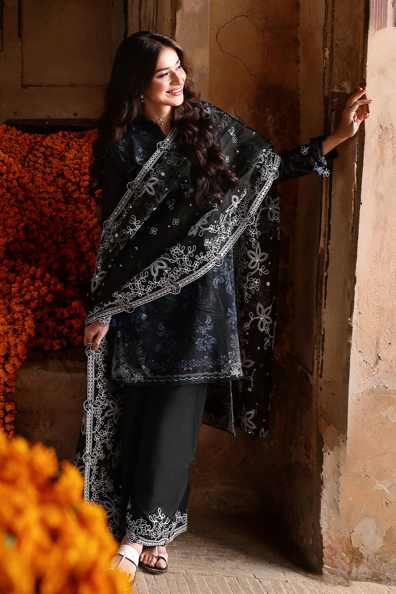 Smiling woman wearing Noir black lawn suit and embroidered dupatta standing next to a rustic beige wall, showcasing the delicate floral embroidery and tassel details on sleeves.