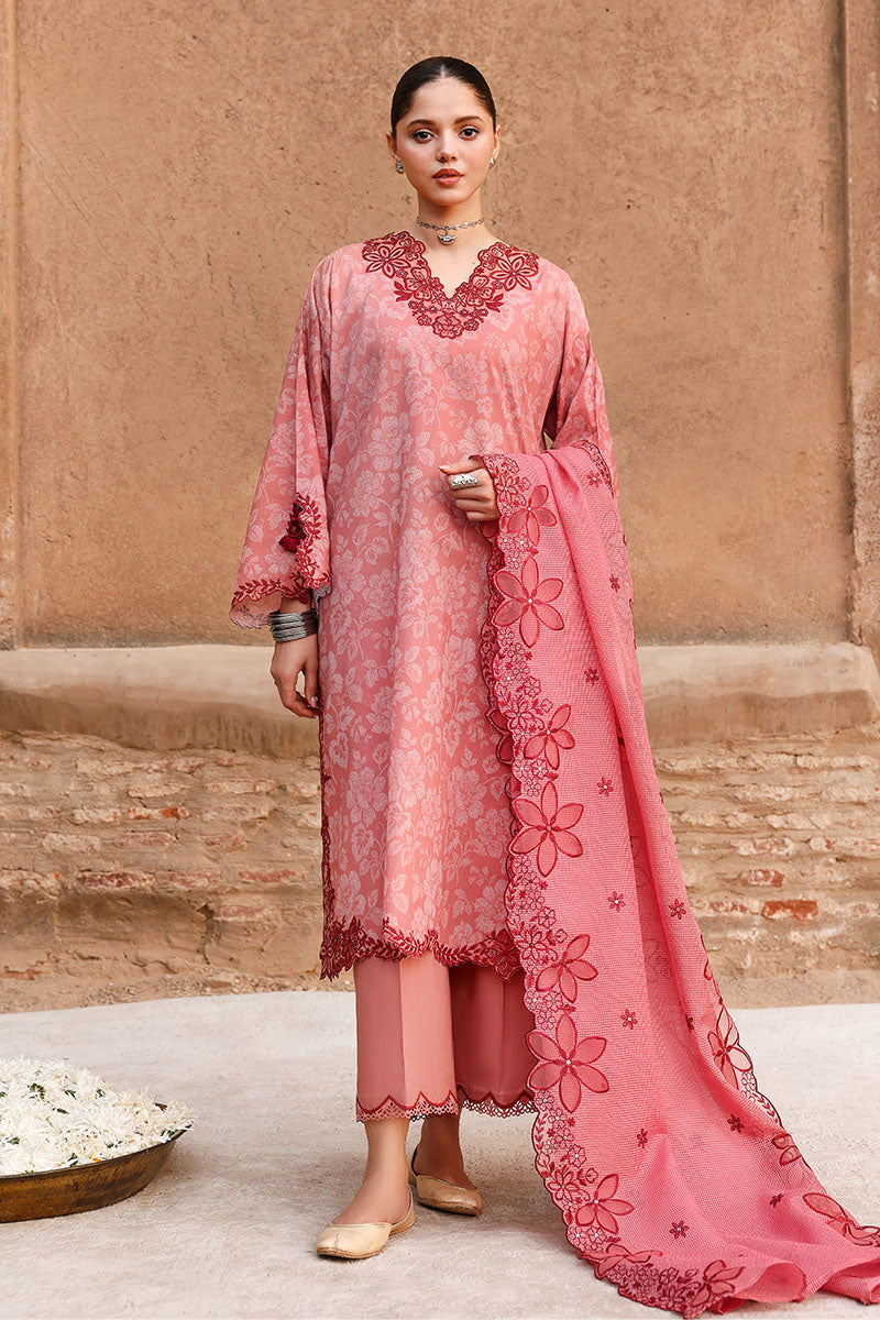 Woman wearing a soft pink embroidered lawn suit with floral patterns and deep red embroidery, standing against a rustic wall.