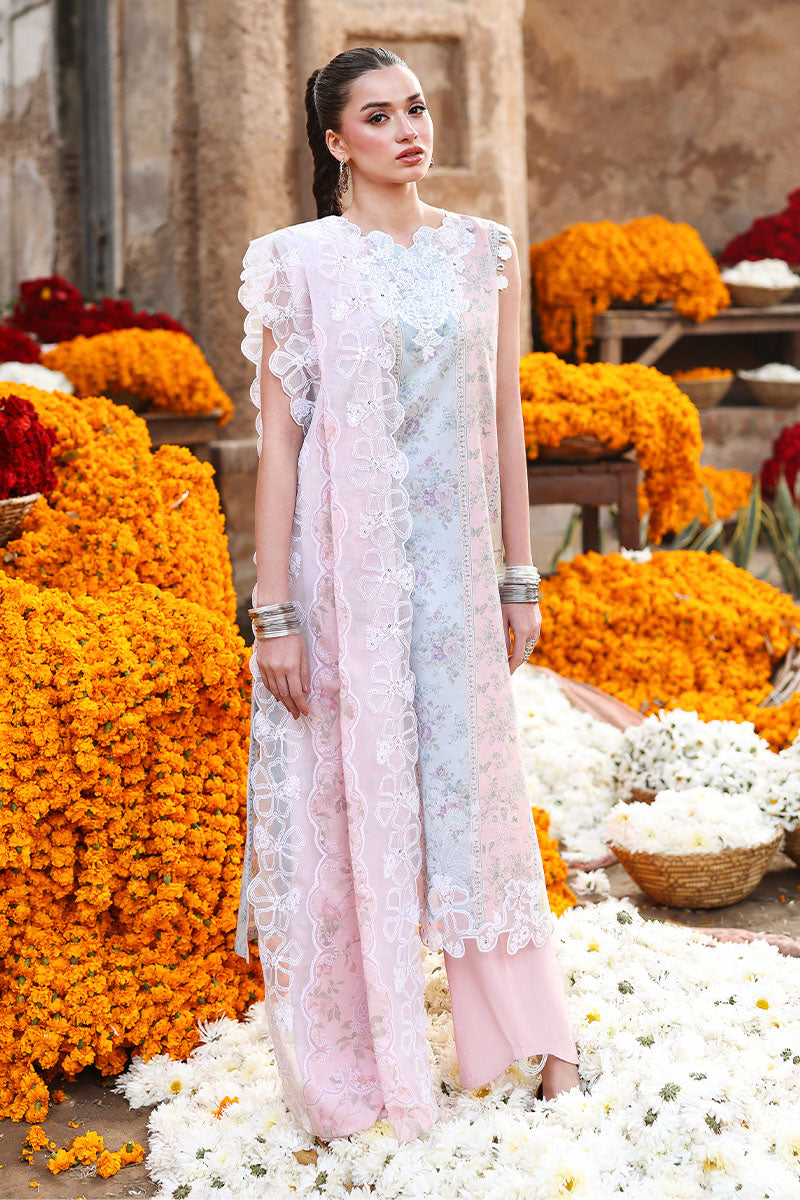 Woman wearing a floral embroidered lawn three-piece suit with light pink trousers and lace-bordered dupatta, standing gracefully amid floral arrangements.