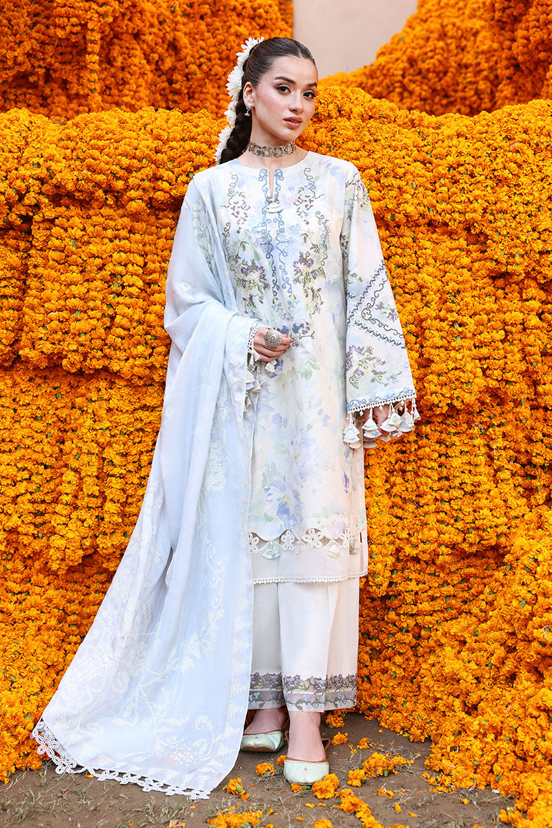 Woman standing wearing Lilac Whisper embroidered lawn suit with pastel floral print and tassel-trimmed sleeves against orange marigold flower backdrop