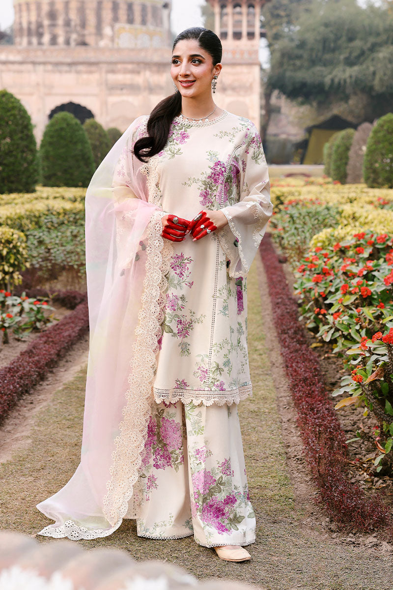 Woman wearing an elegant cream floral lawn suit with purple and green flowers standing in a garden, holding a sheer pink dupatta with lace edges.