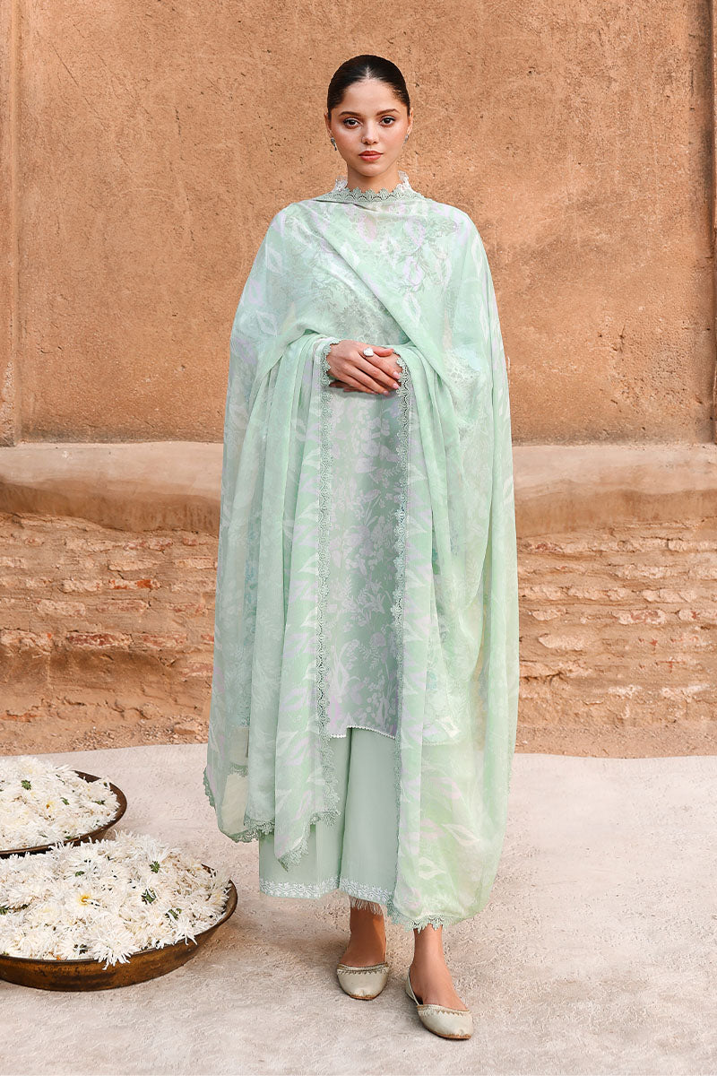 Woman wearing a sage green three-piece lawn suit standing against a textured beige wall, showcasing modest and elegant styling with a printed shirt, matching dyed trousers, and flowing dupatta