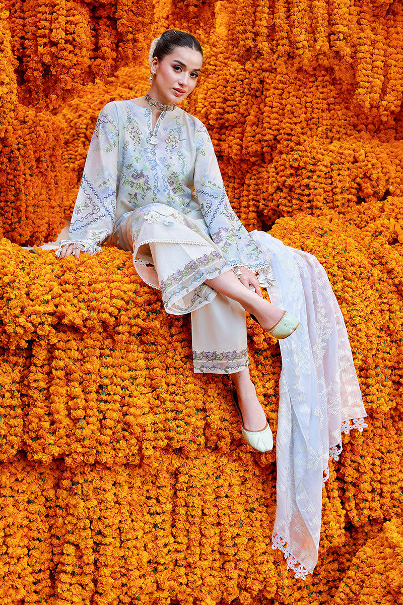 Seated woman in pastel lilac and blue embroidered lawn suit with embroidered trousers and light silk dupatta on marigold flowers