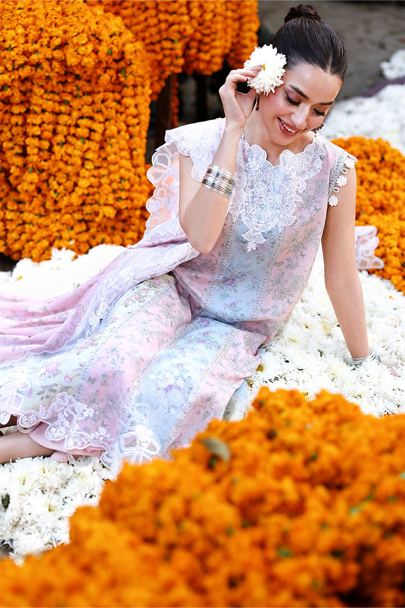 Close-up of a smiling woman seated on white flowers, dressed in a sleeveless pastel floral lawn suit with white embroidery and holding a white flower near her face.