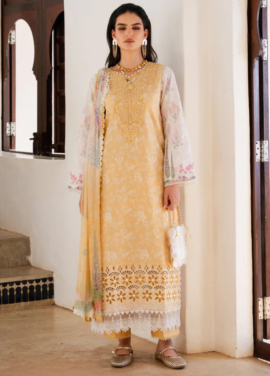 Front view of woman wearing elegant yellow embroidered lawn 3-piece suit with floral patterns and lace hem holding a white handbag indoors