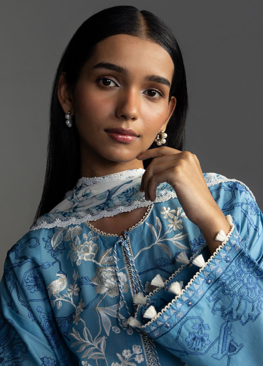 Side close-up of woman's face and upper body highlighting blue floral embroidery on shirt and pearl drop earrings