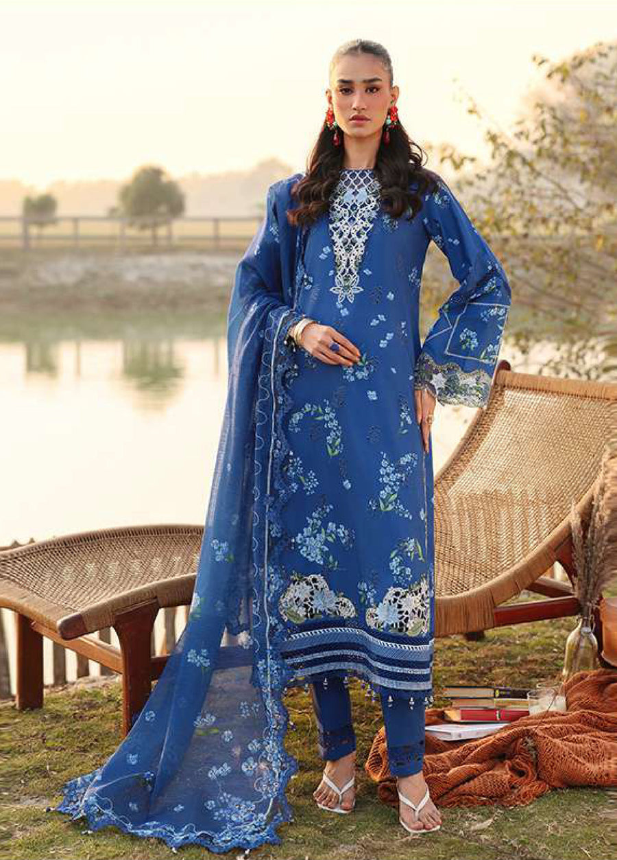Front view of a woman wearing an elegant blue embroidered lawn suit with floral motifs and a sheer dupatta, standing outdoors near a rustic chair