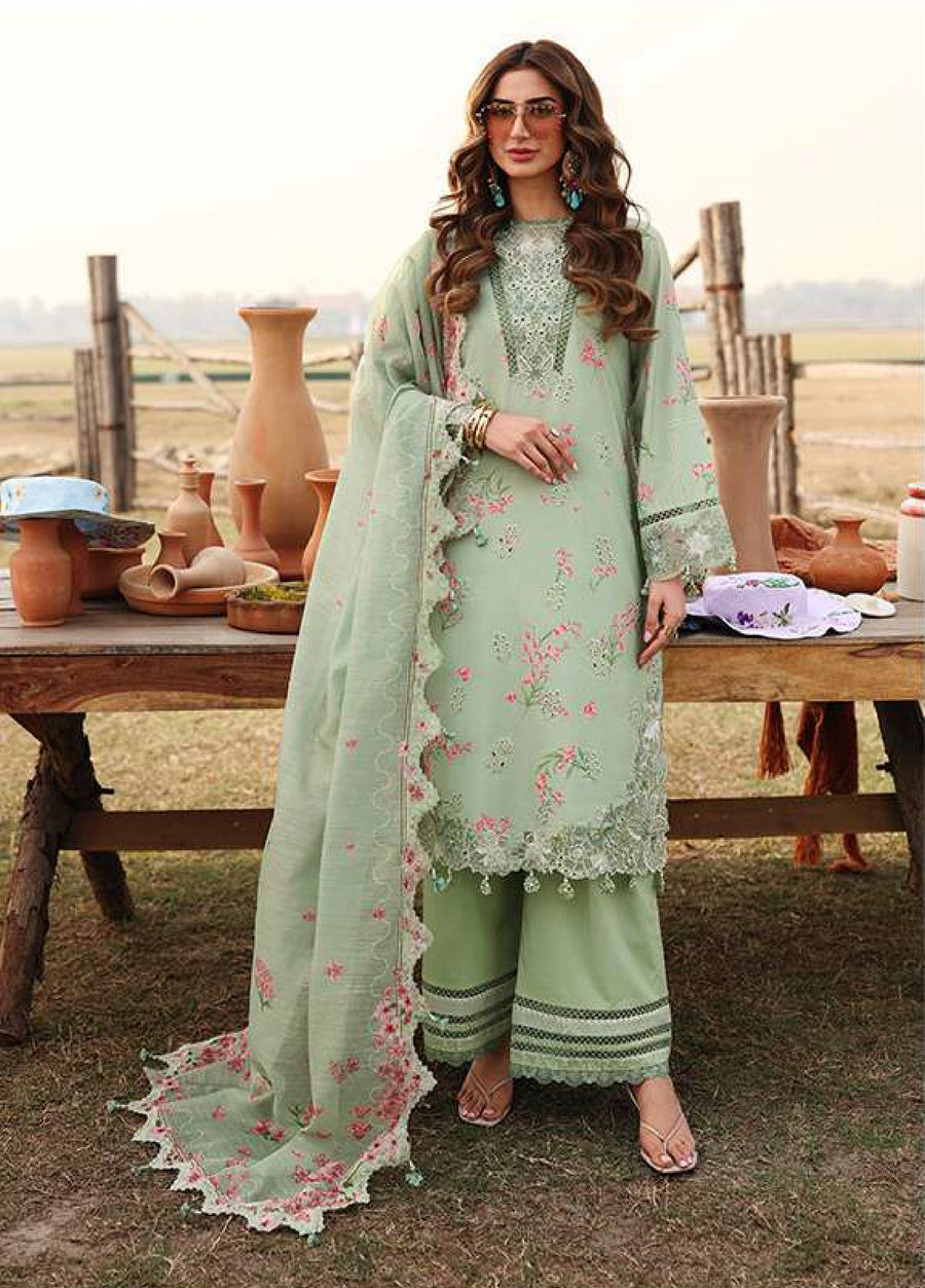 Woman wearing pastel green embroidered lawn suit with floral motifs and matching dupatta standing outdoors by a wooden table with pottery.