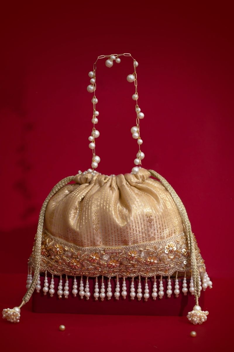 Golden potli bag with embroidered floral pattern and pearl tassels, featuring a pearl-adorned chain handle on a red background