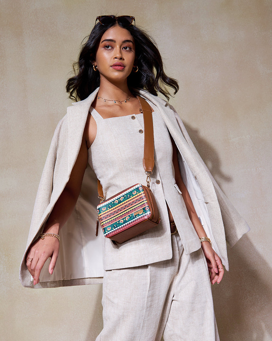 Woman wearing beige linen outfit with beige jacket draped over shoulders, accessorized with a colorful Jaipur Sling crossbody bag with brown leather strap, featuring multi-colored embroidered stripes and floral patterns.