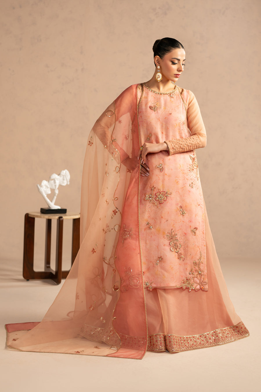 Model holding sheer embroidered peach dupatta with floral and gold detailing, wearing peach embroidered long shirt and sharara
