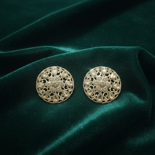 Pair of round gold filigree stud earrings with teardrop and circular crystal design resting on dark green velvet fabric