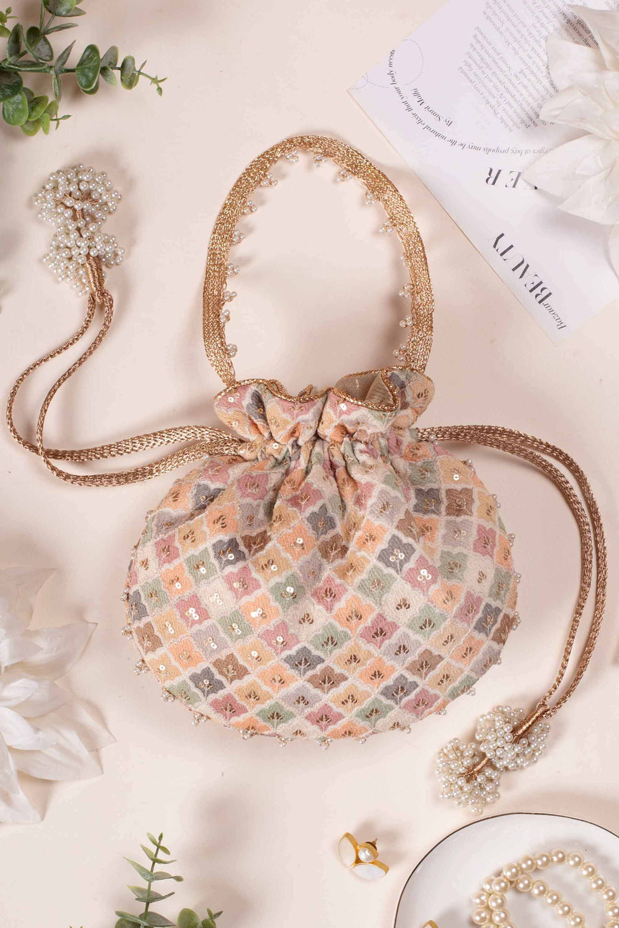 Pastel embroidered potli bag with gold braided drawstrings and pearl tassel embellishments on a soft beige background surrounded by pearl jewelry and greenery