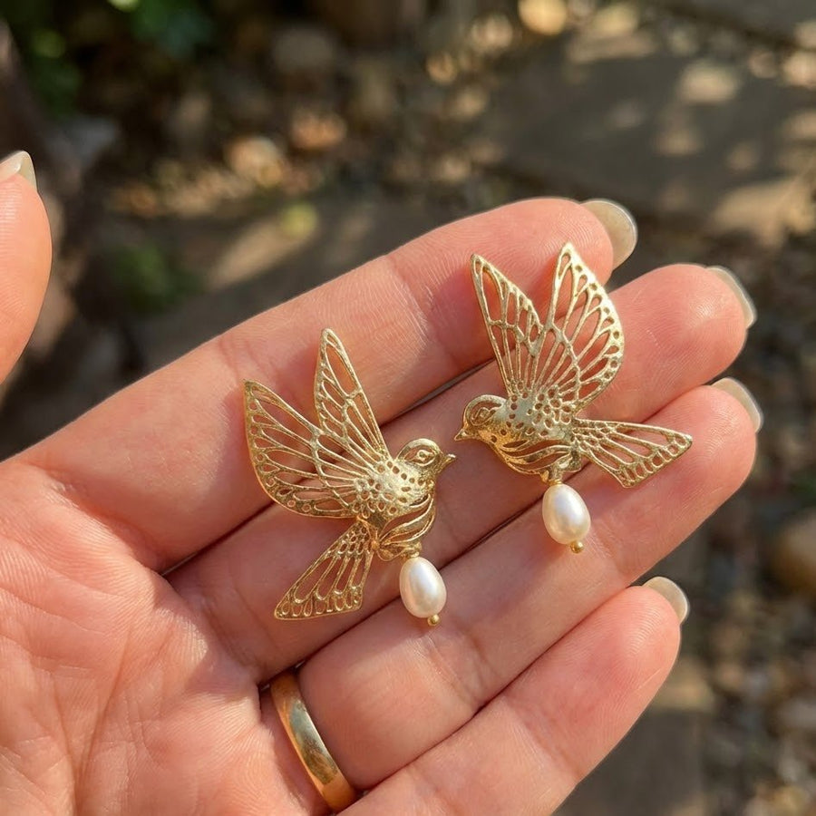 Gold bird pearl drop earrings resting on an open hand with natural outdoor background
