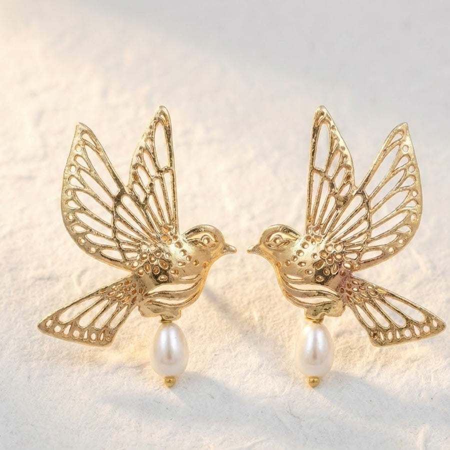 Close-up of gold bird earrings with intricate cut-out wing design and pearl drops on a textured neutral surface