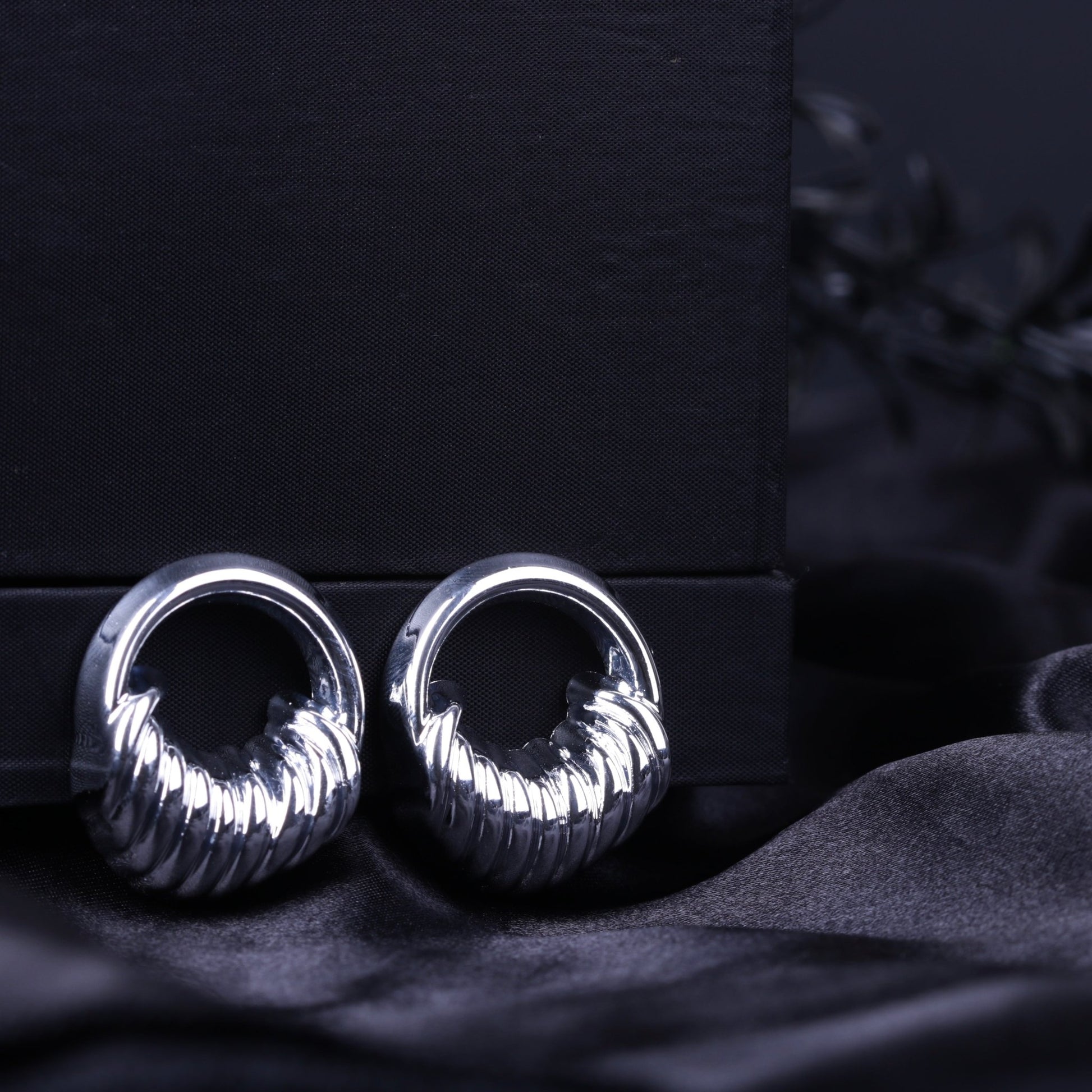 Silver sculpted textured hoop earrings with ribbed detailing on a black fabric background