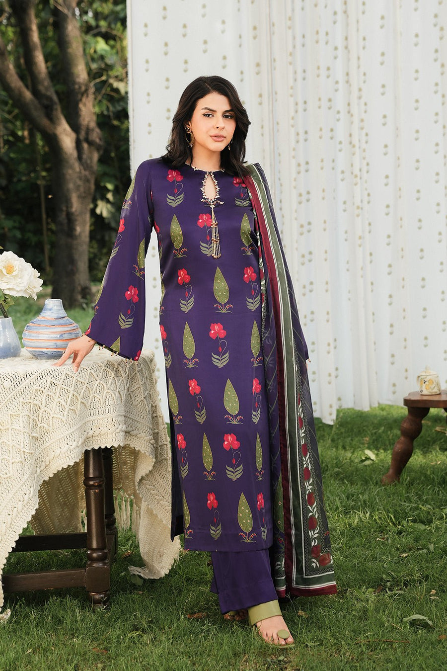 Woman wearing a purple silk floral kurta set with red and green motifs, standing next to a table with a crochet cloth outdoors