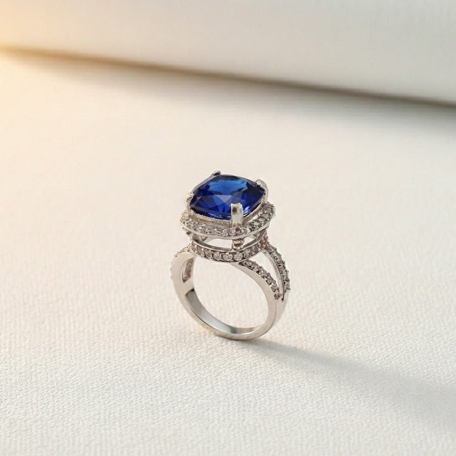 Dark blue cushion-cut gemstone ring with double pavé halo and split silver-tone band on light background
