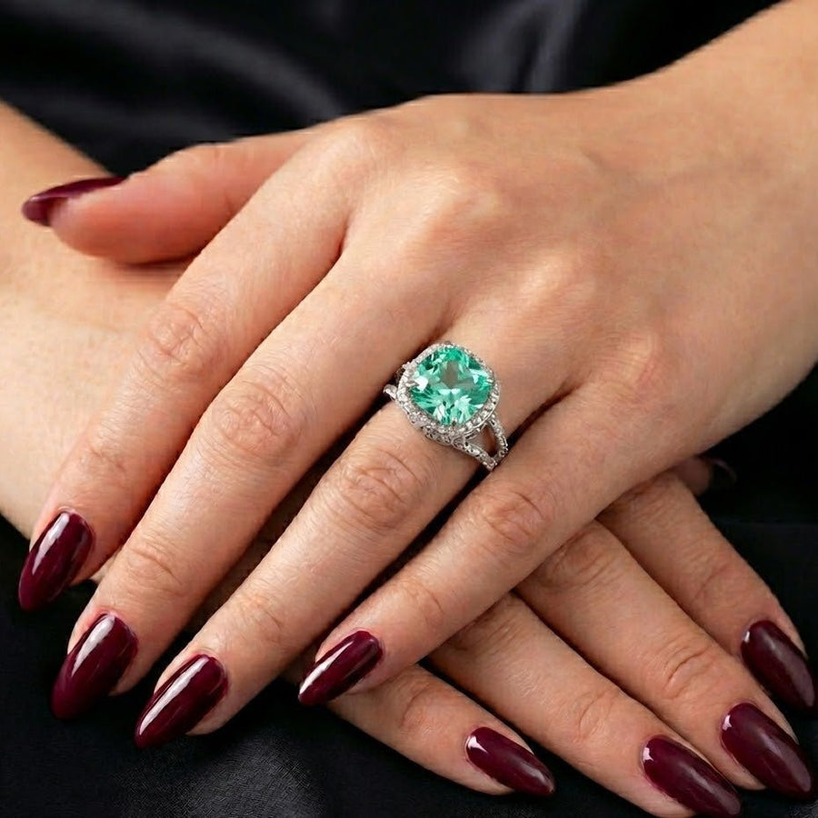 Women's hand displaying cushion-cut green gemstone pavé ring with dark red manicure