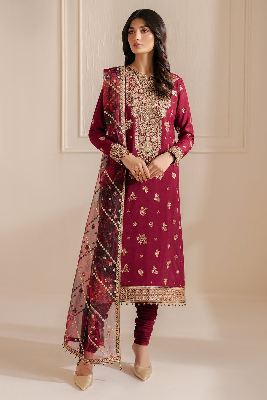 Frontal view of a woman modeling a rich maroon embroidered formal suit with detailed gold embroidery on the chest and cuffs, complemented by a transparent organza dupatta with gold motifs, standing poised in neutral setting.