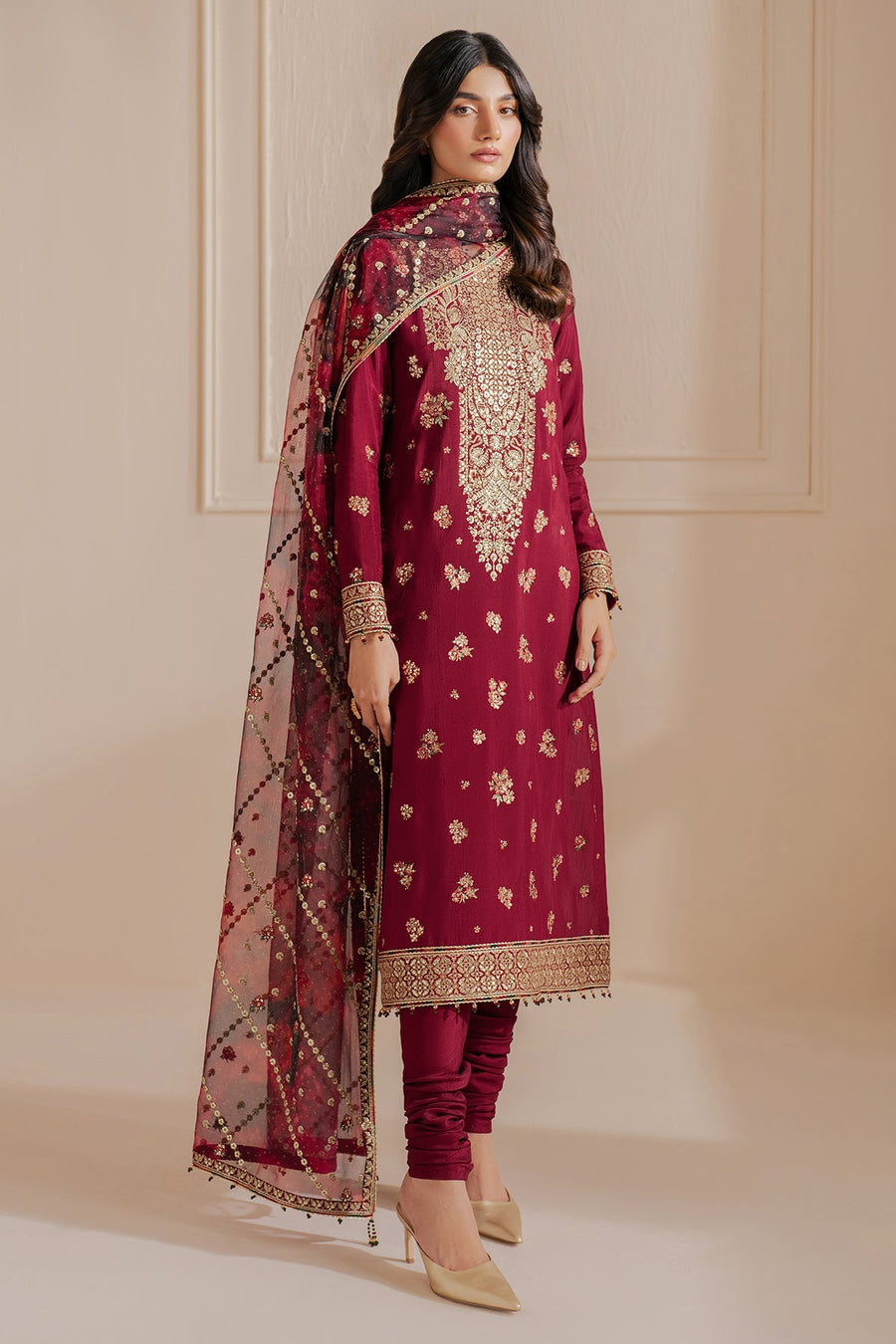 Woman wearing a maroon embroidered formal suit with gold floral patterns, paired with matching churidar pants and a sheer organza dupatta with gold bead embellishments, standing against a neutral background.