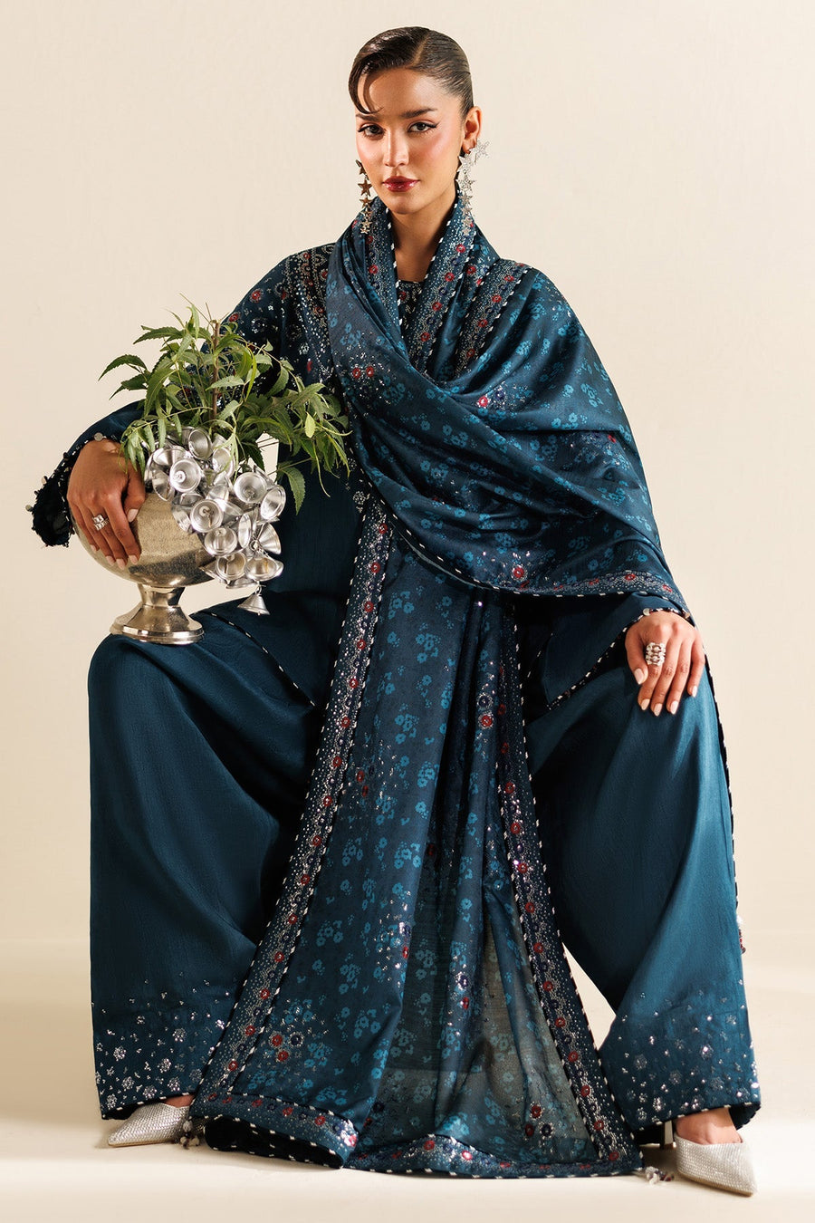 Model seated wearing teal embroidered suit with intricate floral embroidery and holding a decorative silver bowl with greenery.