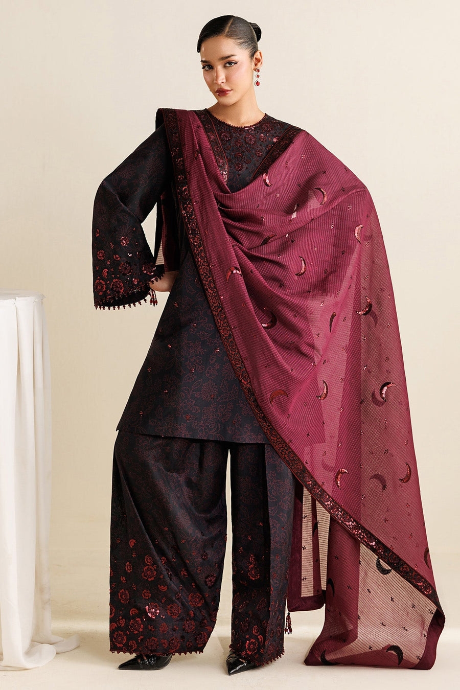 Model gazing downwards with maroon dupatta draped over head and shoulder, highlighting intricate sequin embroidery and floral motifs on black fabric.