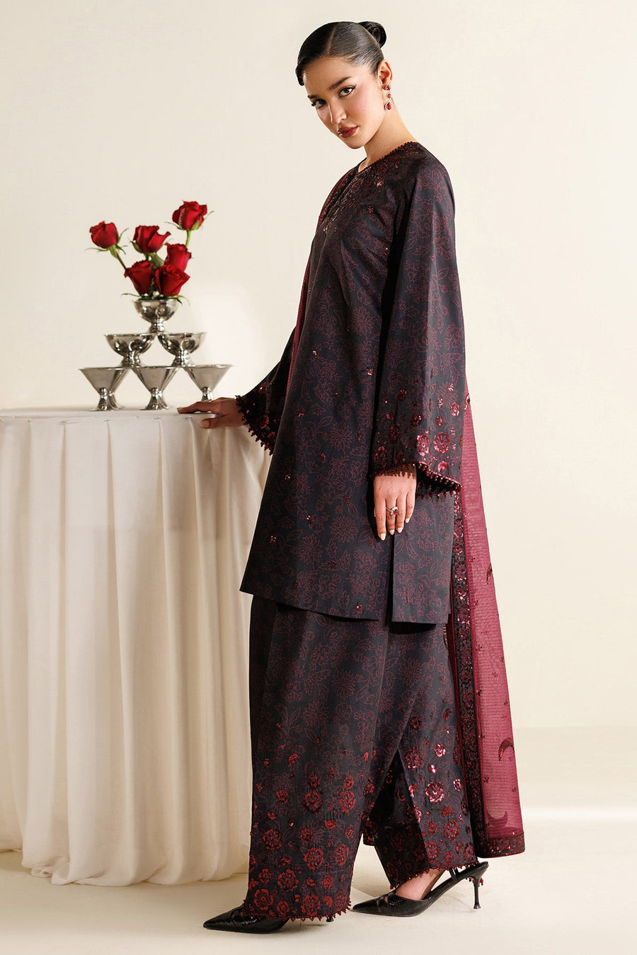 Side view of model displaying black kameez with subtle floral print and sequins, paired with black wide trousers accented with maroon embroidery and bead trim.