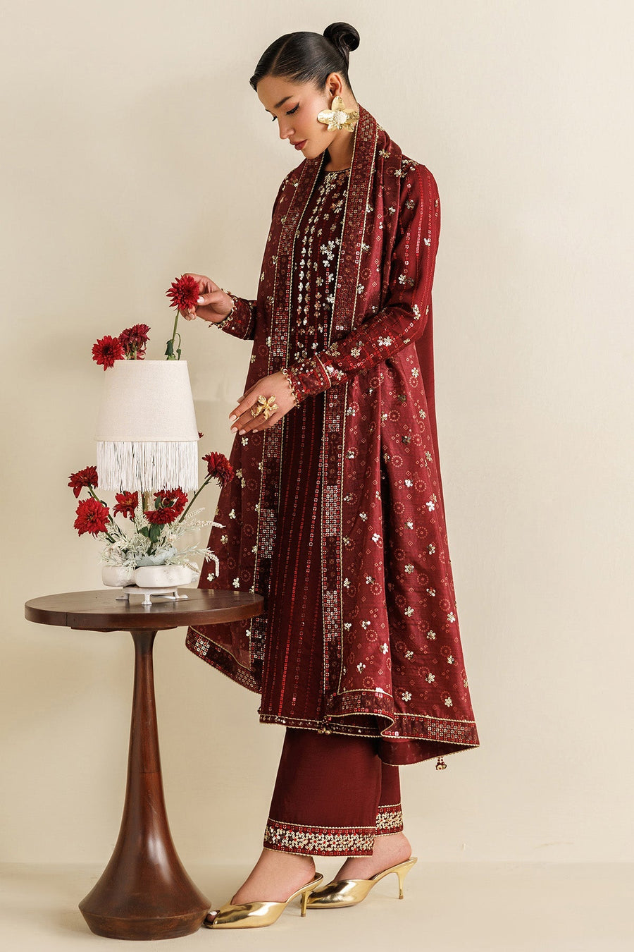 Side view of woman wearing maroon embroidered suit and holding matching dupatta by side table with red flowers