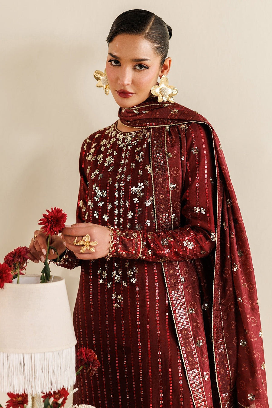 Woman sitting on chair wearing maroon embroidered suit and holding matching dupatta draped over shoulders