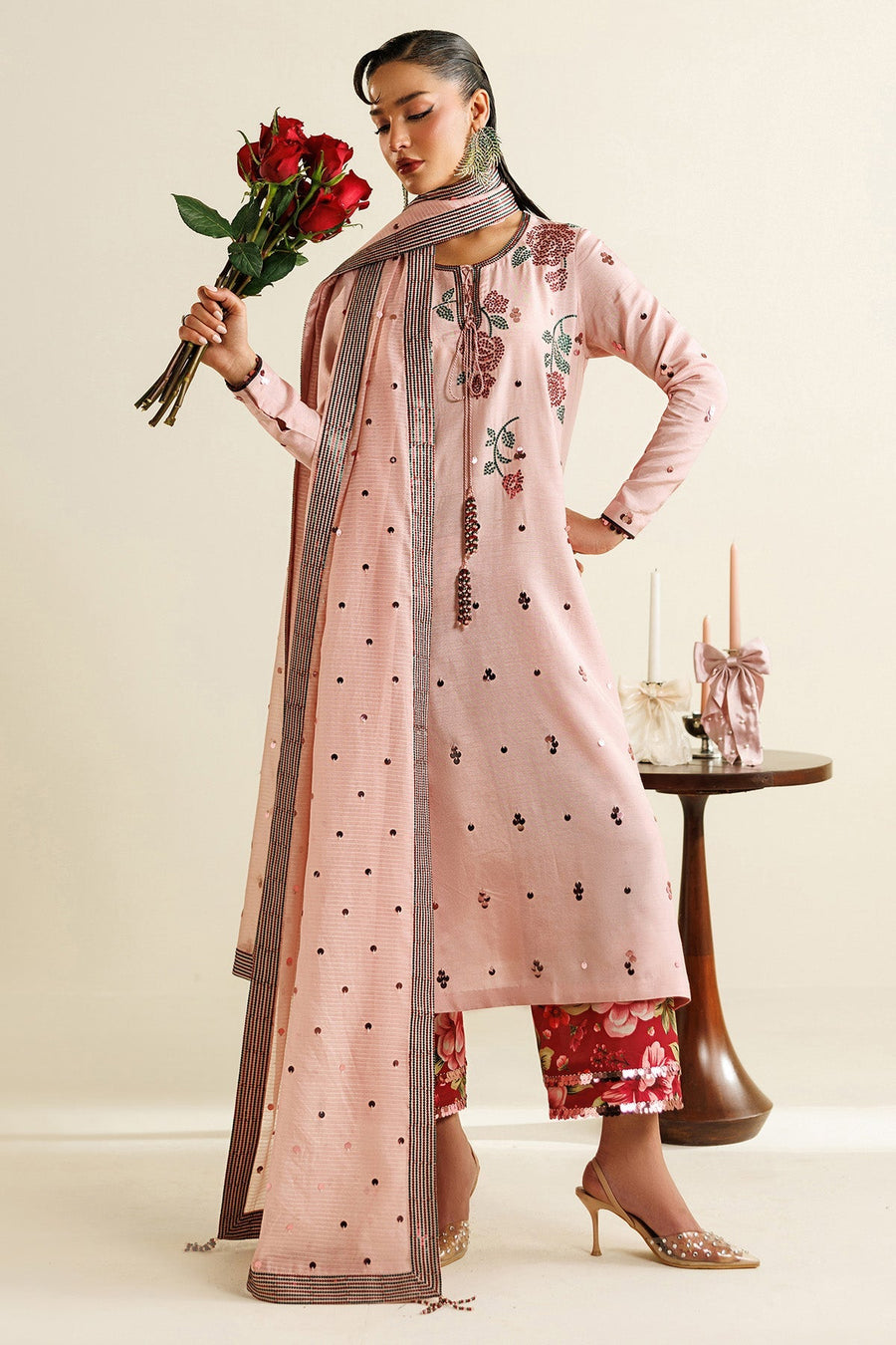 Woman holding bouquet of red roses wearing pink embroidered long shirt with sequin details, red floral pants and matching embellished dupatta