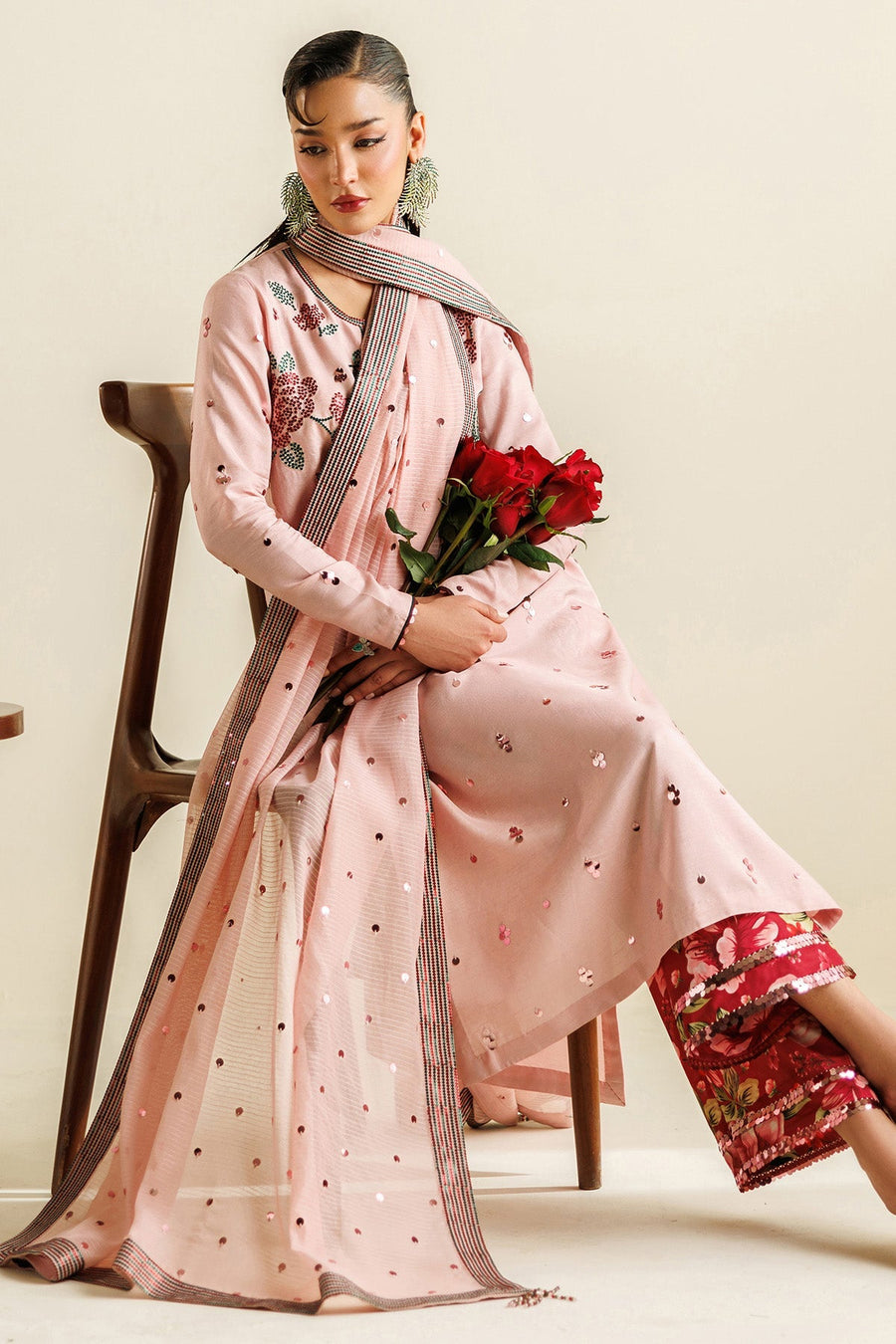 Woman sitting holding red roses, wearing pink embroidered long suit with floral accents, sequin details and matching dupatta