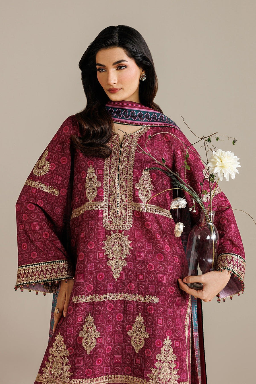 Close-up of model wearing deep magenta embroidered unstitched suit with intricate golden embroidery on neckline and sleeves holding a glass vase with white flowers.
