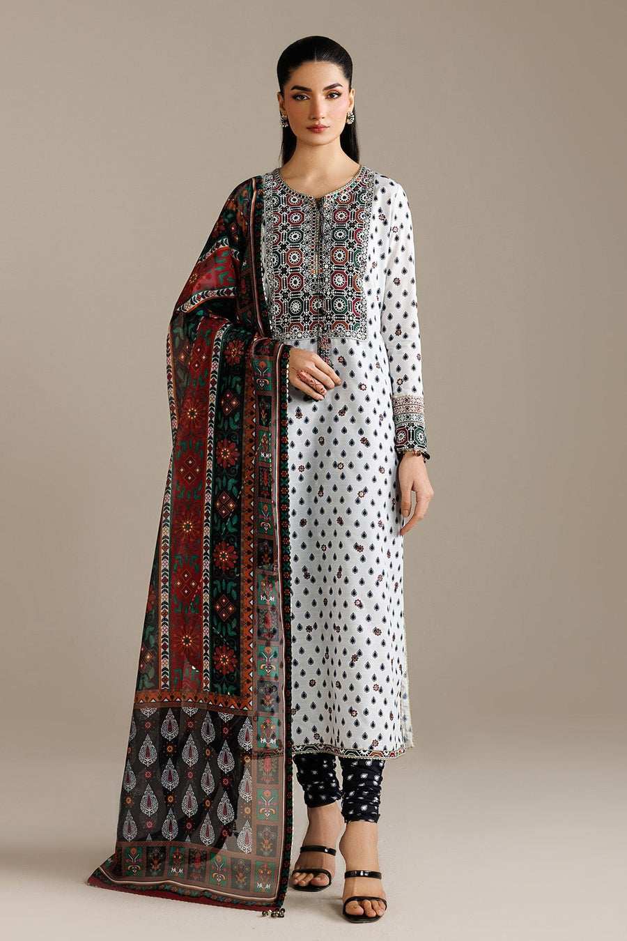 Seated woman wearing embroidered white lawn kurta with detailed neckline and cuffs, styled with printed dupatta and black printed trousers.