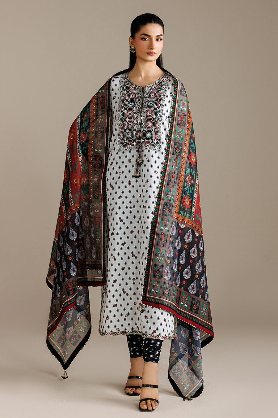 Elegant woman seated on block, styling embroidered white lawn suit with detailed embroidery, paired with printed trousers and a vibrant dupatta draped over one shoulder.