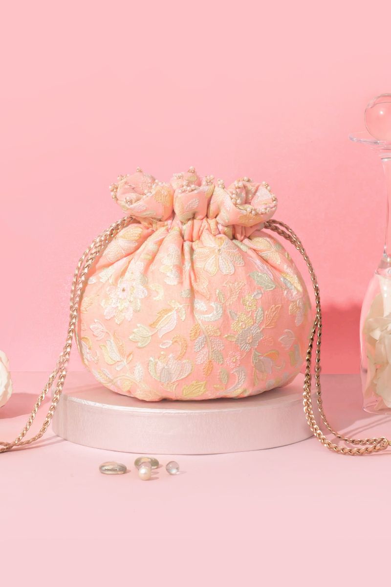 Round peach-colored potli bag with intricate floral embroidery and pearl embellishments, featuring a gold chain strap on a pink background