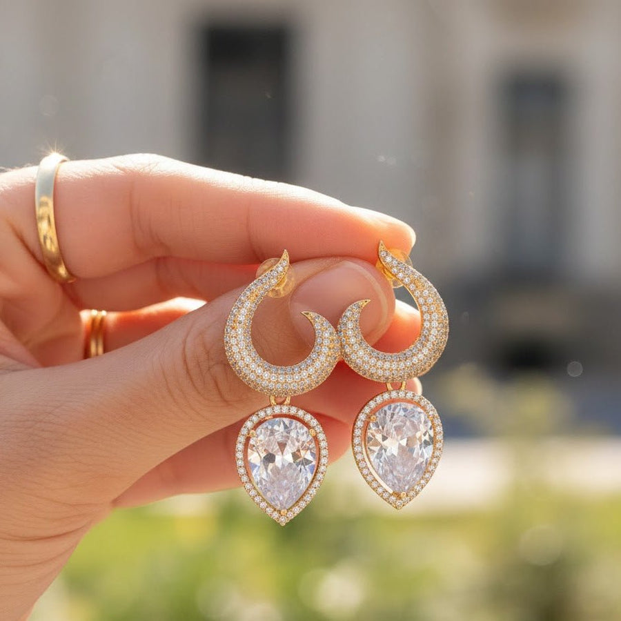 Hand holding gold-plated earrings with clear teardrop gemstones against outdoor background