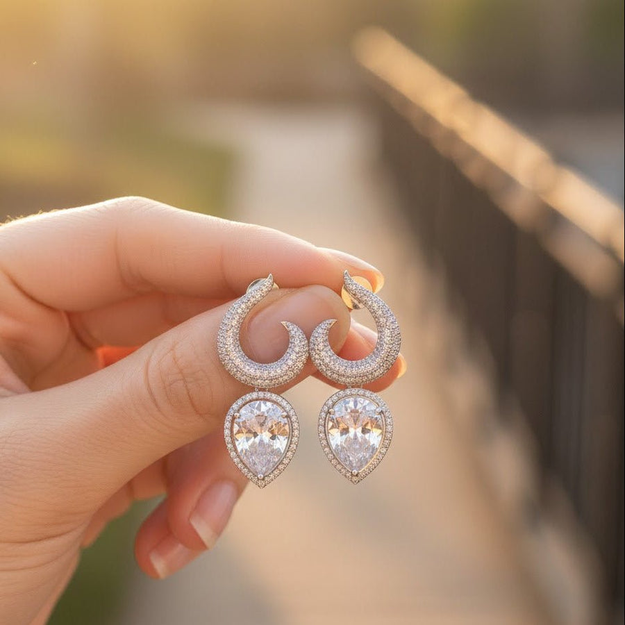 Hand holding silver-plated earrings with clear teardrop gemstones against blurred outdoor background